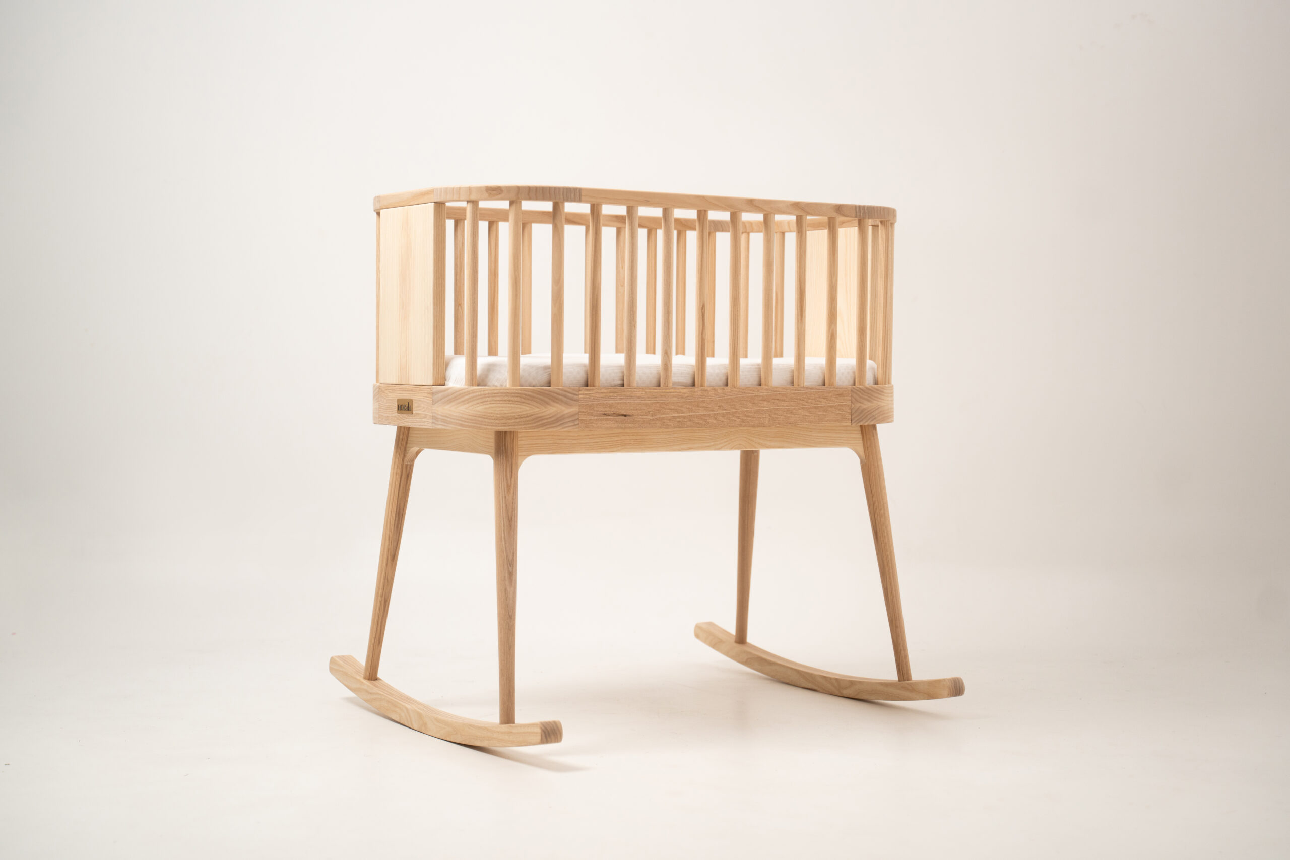 Minimalistyczna drewniana kołyska Norah w kolorze jasnego jesionu na jednolitym tle.